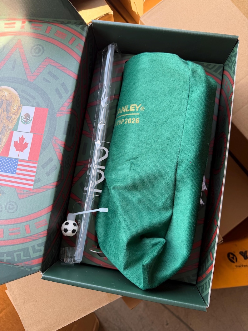 Op de afbeelding: Een groene stoffen hoes met de gouden tekst STANLEY CUP 2026, een doorzichtig plastic rietje met een kleine voetbalbedel en een groene doos met de vlaggen van Mexico, Canada en de Verenigde Staten op de binnenzijde van het deksel.