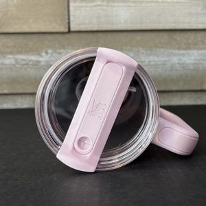 May include: A clear, round lid with a light pink flip-top closure. The closure features a small, embossed design and a circular button. The lid is designed for a tumbler or travel mug, offering a secure seal.