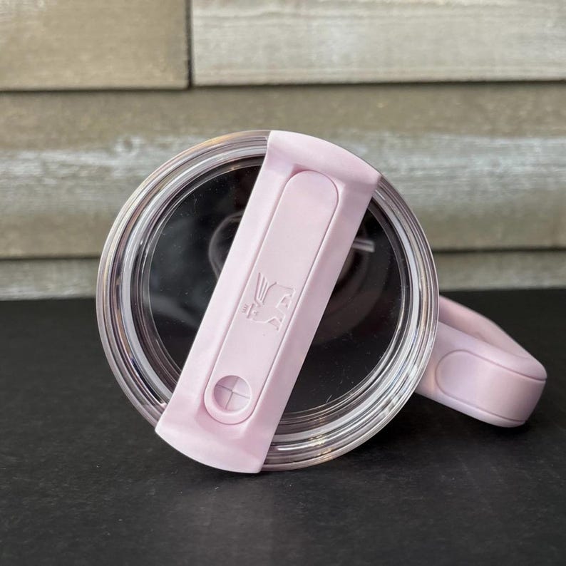 May include: A clear, round lid with a light pink flip-top closure. The closure features a small, embossed design and a circular button. The lid is designed for a tumbler or travel mug, offering a secure seal.