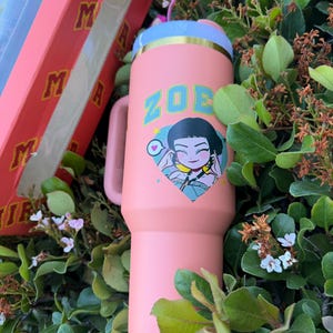 Puede incluir: Un vaso rosa con asa, con el nombre "ZOE" en letras turquesas y un gr&aacute;fico de dibujos animados. El vaso tiene un borde azul claro y dorado y un asa rosa a juego. Se ve una caja rosa con los logotipos "MIRA" y "M".