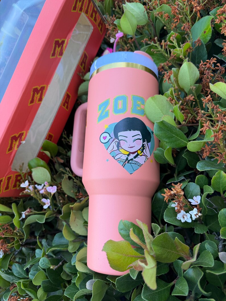 Puede incluir: Un vaso rosa con asa, con el nombre "ZOE" en letras turquesas y un gr&aacute;fico de dibujos animados. El vaso tiene un borde azul claro y dorado y un asa rosa a juego. Se ve una caja rosa con los logotipos "MIRA" y "M".