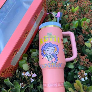 Puede incluir: Un vaso Stanley rosa con asa, pajita y tapa, junto a su caja a juego. El vaso presenta un gr&aacute;fico de dibujos animados y el texto "BUM HUNTRX" y "OP DEMON HUNTR". La caja es rosa con el logotipo de Stanley.