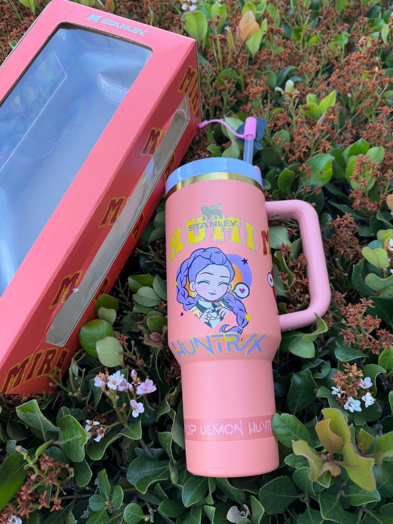 Puede incluir: Un vaso Stanley rosa con asa, pajita y tapa, junto a su caja a juego. El vaso presenta un gr&aacute;fico de dibujos animados y el texto "BUM HUNTRX" y "OP DEMON HUNTR". La caja es rosa con el logotipo de Stanley.