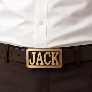 Peut inclure: Une ceinture en cuir marron foncé dotée d'une boucle rectangulaire en métal doré. La boucle présente un design découpé affichant le nom JACK en lettres capitales grasses. La ceinture est portée sur un pantalon marron et une chemise blanche.