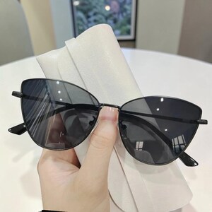 Op de afbeelding: Zwarte cat-eye zonnebril met een dun, donker metalen montuur. De glazen zijn donker en de zonnebril wordt in een witte etui gehouden. De zonnebril heeft een strak, modern design.