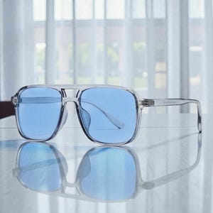 May include: Clear-framed sunglasses with blue lenses. The square-shaped frames have a double bridge and a subtle, transparent design. The sunglasses are reflected on a smooth surface, suggesting a modern aesthetic.