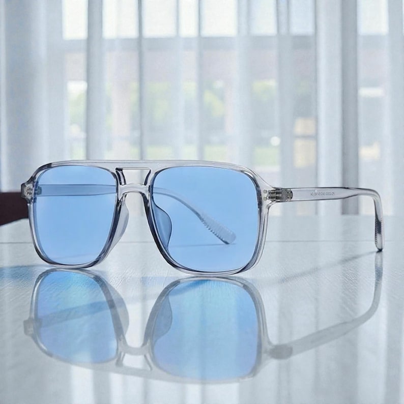 May include: Clear-framed sunglasses with blue lenses. The square-shaped frames have a double bridge and a subtle, transparent design. The sunglasses are reflected on a smooth surface, suggesting a modern aesthetic.