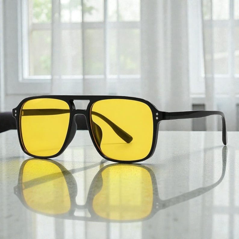 May include: Black-framed sunglasses with yellow lenses. The sunglasses have a square shape and a double bridge design. The lenses are a bright yellow color, and the frames are black. The sunglasses are reflected on a shiny surface.