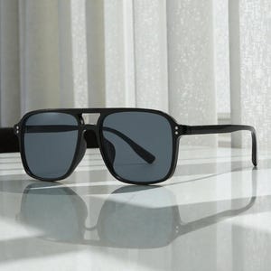 May include: Black square-framed sunglasses with dark gray lenses. The sunglasses have a double bridge and a sleek, modern design. The frames are thick and the temples are straight.