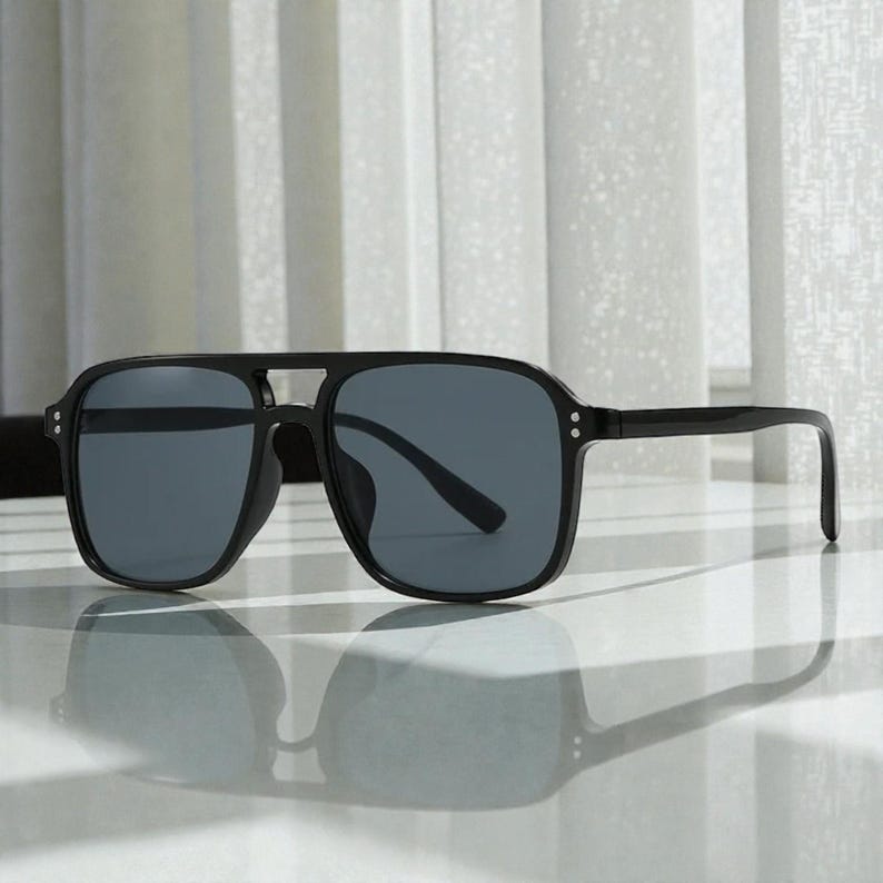 May include: Black square-framed sunglasses with dark gray lenses. The sunglasses have a double bridge and a sleek, modern design. The frames are thick and the temples are straight.