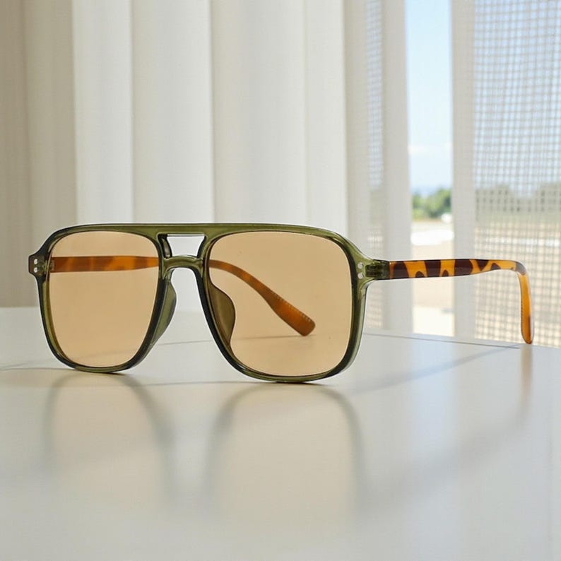 May include: Olive green square-framed sunglasses with amber-colored lenses. The temples feature a tortoise shell pattern. The sunglasses are displayed on a white surface, with a window and sheer curtains in the background.