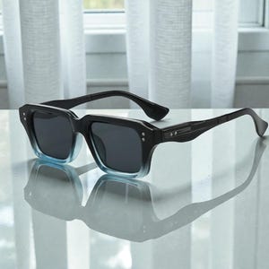 May include: Black sunglasses with a square frame and gradient blue tint at the bottom. The sunglasses have a sleek, modern design with dark lenses and a subtle reflection on a light-colored surface. The arms are black and feature small silver accents.