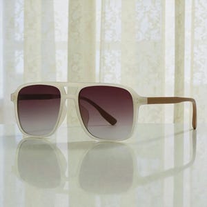 May include: Square-framed sunglasses with a light cream-colored frame and brown gradient lenses. The temples are a light brown color. The sunglasses are sitting on a reflective surface.