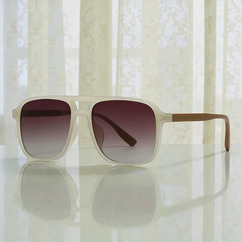 May include: Square-framed sunglasses with a light cream-colored frame and brown gradient lenses. The temples are a light brown color. The sunglasses are sitting on a reflective surface.