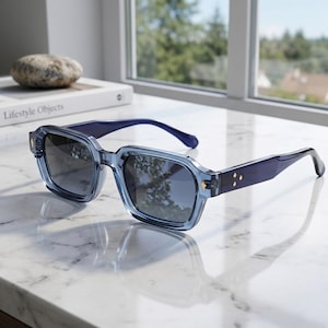 May include: Blue square-framed sunglasses with dark lenses. The sunglasses have a translucent blue frame and dark blue arms. Gold-coloured accents are visible on the frame. The sunglasses are resting on a white marble surface.