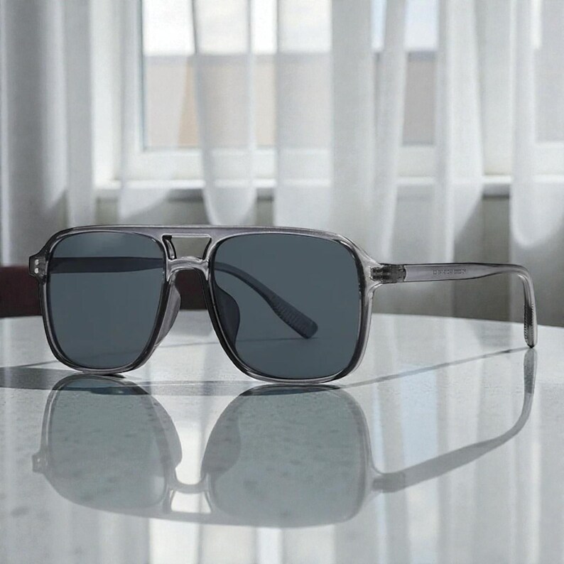 May include: Gray sunglasses with dark lenses. The sunglasses have a square frame and a double bridge. The temples are thin and transparent. The sunglasses are reflected on a shiny surface.