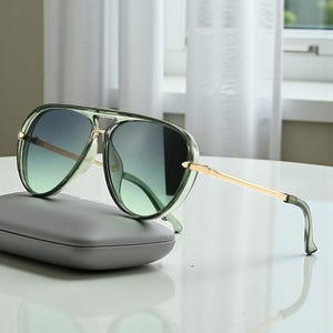 以下が含まれることがあります： グラデーションレンズとゴールドカラーのアクセントが付いたグリーンのアビエーターサングラス。サングラスはグレーのケースの上に置かれています。レンズは緑から濃い色合いに変化します。