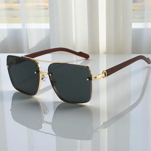 May include: Square-framed sunglasses with a gold metal frame and dark lenses. The temples are a dark wood grain. The sunglasses are reflected on a white surface.