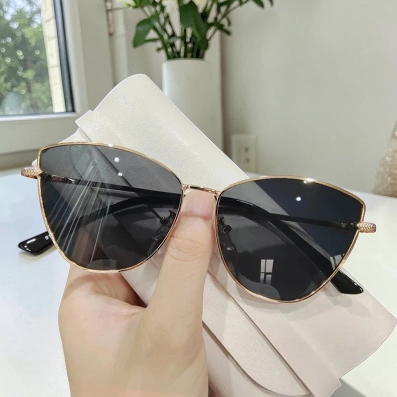 Op de afbeelding: Goudkleurige zonnebril met donkere glazen. De zonnebril in cat-eye-vorm wordt tegen een witte achtergrond gehouden. De monturen hebben een delicaat ontwerp bij de slapen. De glazen zijn donker en reflecterend.
