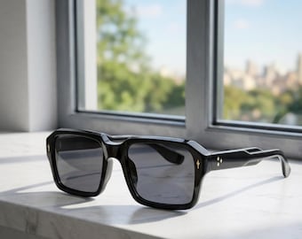 May include: Black square-framed sunglasses with dark lenses. The sunglasses have gold-colored accents on the temples and the frame. The sunglasses are sitting on a white surface in front of a window.