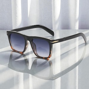 May include: Square-framed sunglasses with black frames and gradient blue lenses. The sunglasses feature a tortoiseshell pattern on the lower frame and a gold-coloured detail on the temples. The lenses transition from dark blue to a lighter shade.
