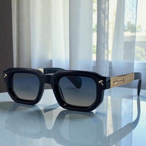 May include: Black sunglasses with a geometric frame and gradient lenses. The temples feature gold-colored accents and the letter "T". The sunglasses are displayed on a reflective surface.