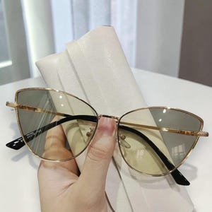 Op de afbeelding: Goudkleurige cat-eye zonnebril met zwarte pootjes en getinte glazen. De bril wordt voor een witte etui gehouden. De monturen hebben een delicaat, stijlvol ontwerp, geschikt voor mode en oogbescherming.