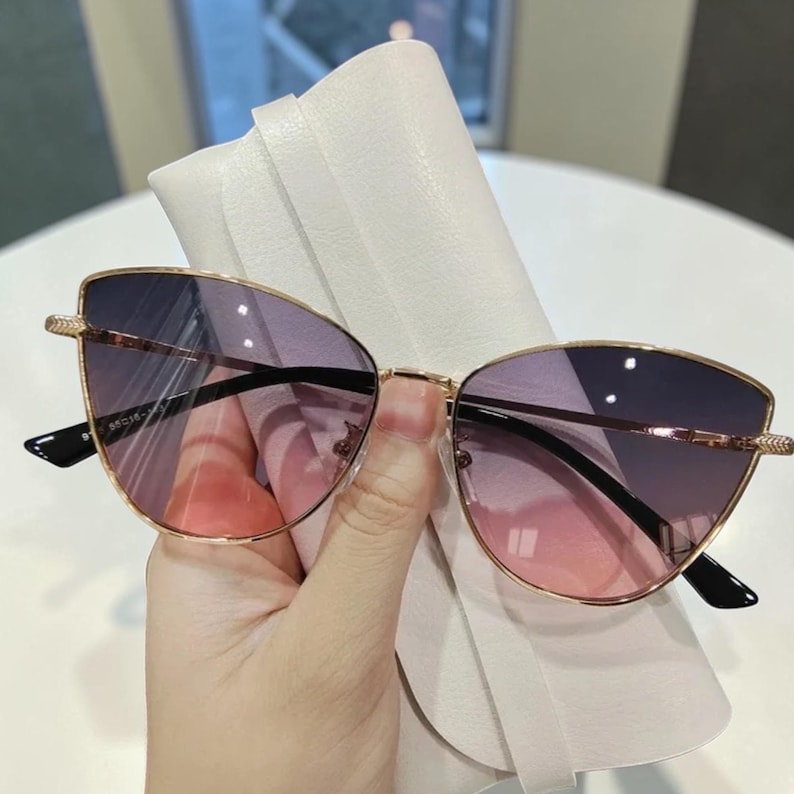 Op de afbeelding: Cat-eye zonnebril met een goudkleurig metalen montuur en verloopglazen die overgaan van roze naar paars. De zonnebril wordt in een witte etui gehouden. De tekst "MCM" is zichtbaar op de linker poot.
