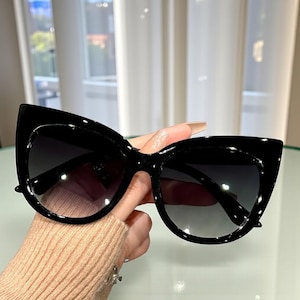 Puede incluir: Gafas de sol negras de ojo de gato con lentes degradadas. Las gafas tienen un acabado brillante y una montura gruesa. Las lentes hacen una transición de oscuro en la parte superior a más claro en la parte inferior. Las gafas son sostenidas por una persona.