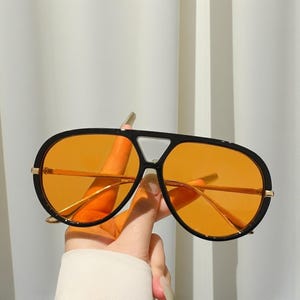May include: Black aviator sunglasses with orange tinted lenses. The sunglasses have a gold-colored metal frame and a black plastic frame. The sunglasses are held by a hand.