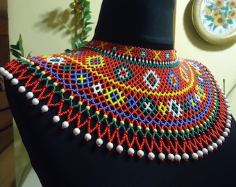 May include: A colorful beaded necklace with a geometric pattern in red, blue, green, yellow, and white. The necklace is made of small beads and has a fringe at the bottom.