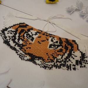 May include: A beaded necklace with a tiger face design. The necklace is made of white, black, and gold beads. The beads are arranged in a fringe pattern around the tiger face.