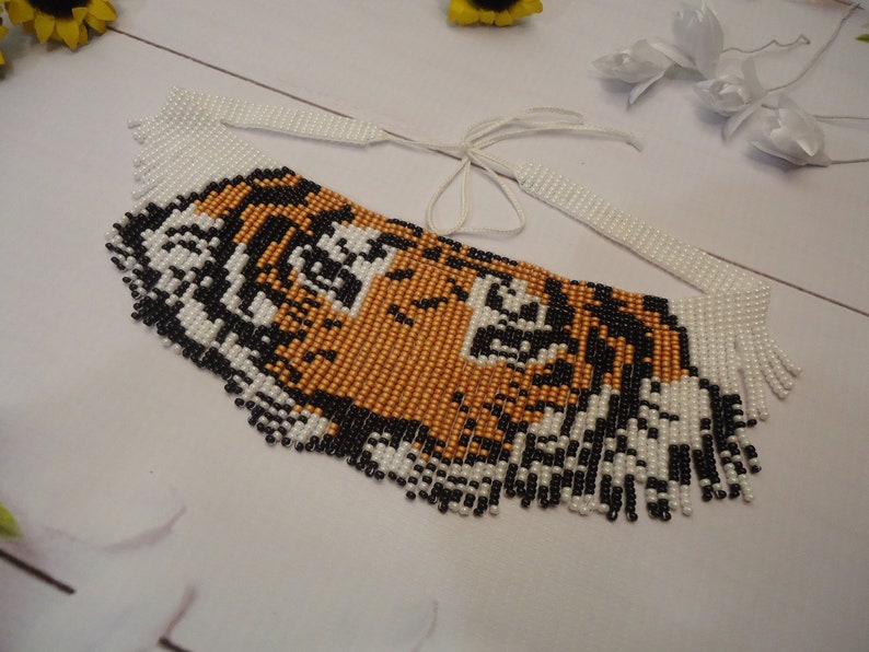 May include: A beaded necklace with a tiger face design. The necklace is made of white, black, and gold beads. The beads are arranged in a fringe pattern around the tiger face.