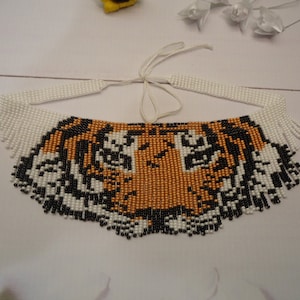 May include: A beaded necklace with a tiger face design. The necklace is made with white, black, and orange beads. The tiger face is detailed with black and white beads for the eyes and stripes.