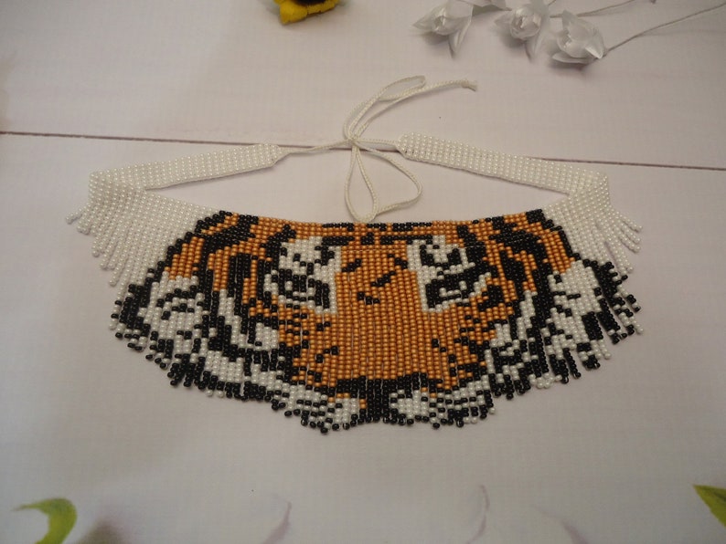 May include: A beaded necklace with a tiger face design. The necklace is made with white, black, and orange beads. The tiger face is detailed with black and white beads for the eyes and stripes.