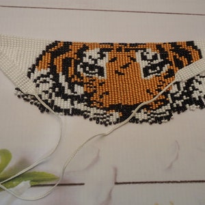 May include: A beaded tiger face necklace with white, black, and orange beads. The necklace has two white cords attached to the bottom.