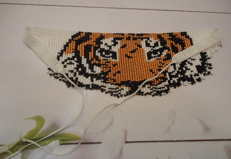 May include: A beaded tiger face necklace with white, black, and orange beads. The necklace has two white cords attached to the bottom.