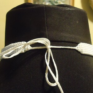May include: White beaded necklace with a white cord tie closure. The necklace is displayed on a black mannequin.