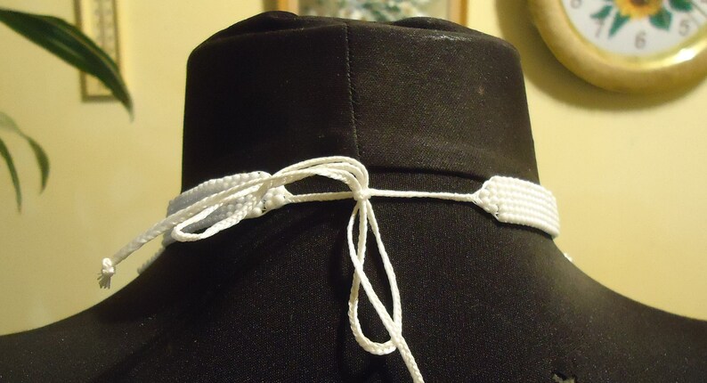 May include: White beaded necklace with a white cord tie closure. The necklace is displayed on a black mannequin.