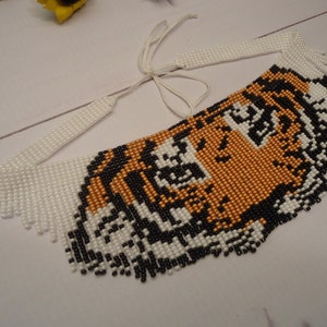 May include: A beaded necklace with a tiger face design. The tiger's face is made of black, white, and orange beads. The necklace has a white fringe around the edges.