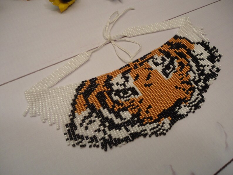 May include: A beaded necklace with a tiger face design. The tiger's face is made of black, white, and orange beads. The necklace has a white fringe around the edges.