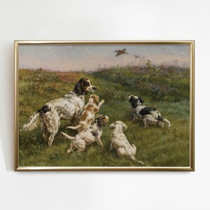 May include: A framed vintage painting depicts a hunting scene with several dogs in a grassy field, with a bird flying overhead. The dogs are various colors, including white, brown, and black. The artwork is framed in a gold-colored frame.