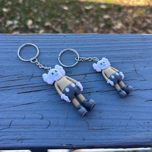 May include: Two keychains featuring a cartoon character with a white head, tan body, and gray shorts. The character has black X's for eyes and is attached to a silver keyring. The keychains are lying on a blue wooden surface.