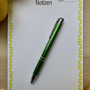 May include: A white notepad with a dotted grid pattern and a yellow lemon border. The top centre features the word Notizen and a line for Datum. A green ballpoint pen with the text Lemonplan.de is placed diagonally across the centre of the page.