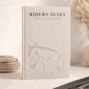 Op de afbeelding: Een beige Riders Diary-dagboek met een paardenschets en de tekst "RIDERS DIARY TRAINING • FORTSCHRITT • GEFÜHLE". Een zwarte leren riem met zilveren gespen staat op de voorgrond. Een touw en gedroogde planten zijn ook zichtbaar.