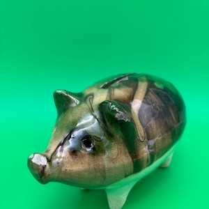 May include: A ceramic piggy bank featuring a glossy, marbled glaze in shades of dark green, brown, and black. The pig-shaped coin bank has a coin slot on its back, pointed ears, a snout, and four short, solid white legs supporting its rounded body.