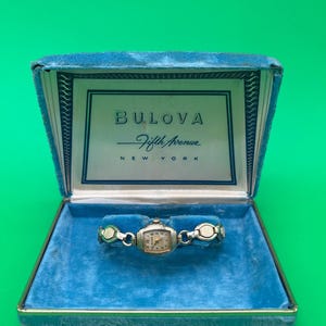 May include: A silver-toned Bulova Fifth Avenue New York wristwatch with a rectangular face and a link bracelet, presented inside a blue velvet-lined display box. The interior lid features the text Bulova Fifth Avenue New York in dark blue.