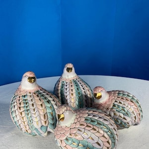 May include: Four ceramic bird figurines with pink, teal, and gold feather detailing. The birds have gold beaks and are arranged on a white surface against a blue backdrop. The figurines are decorative and have a detailed, textured appearance.