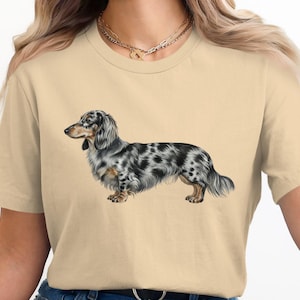 May include: A beige t-shirt with a detailed illustration of a long-haired dachshund dog. The dog has a spotted coat in shades of grey and black. The dog is in profile, showing its distinctive features.