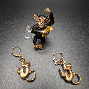 May include: A black monkey brooch with gold-tone accents, clear crystals on the ears and face, and a yellow banana, accompanied by a pair of gold-tone monkey-shaped drop earrings with lever-back closures, featuring a polished metallic finish.